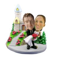 Custom bobble heads depict a handsome groom carrying is sweet heart at in front of a traditional church. The custom bobblehead groom is dressed in a black suit, black tie and big leather shoes. The delighted custom bobbblehead bride is in pink wedding gow